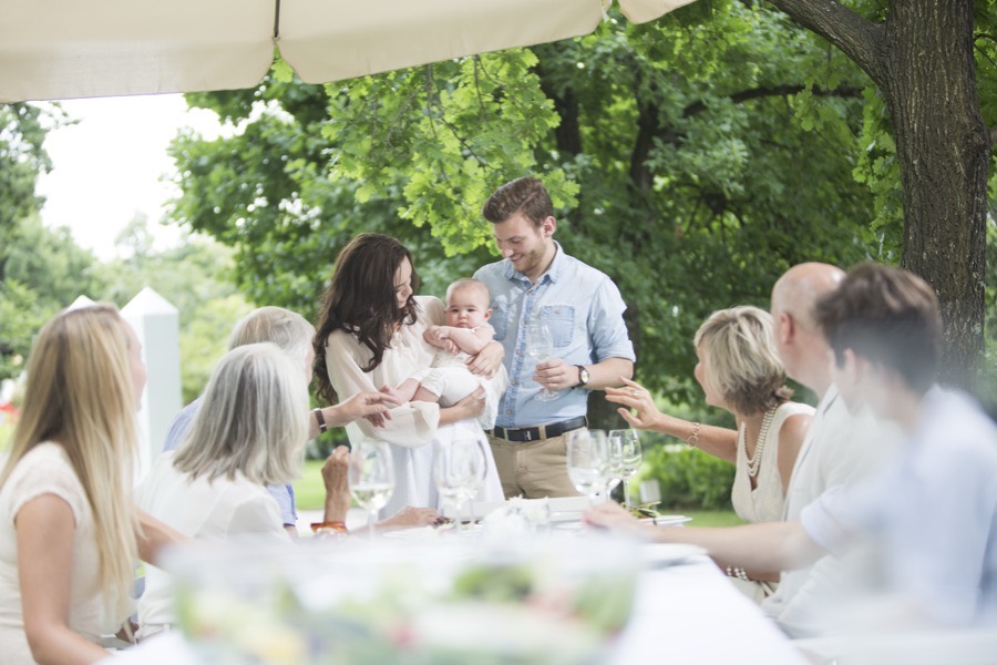 Family gathered around a long table in a garden, meeting a new baby